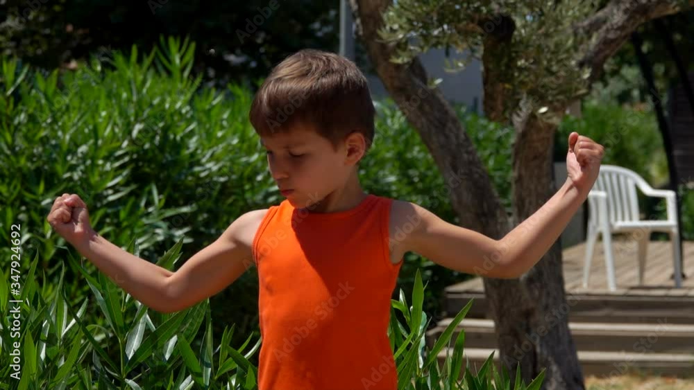 Little cheerful boy in an orang t-shirt is showing his biceps muscle outdoors Little cheerful boy in an orang t-shirt is showing his biceps muscle outdoors