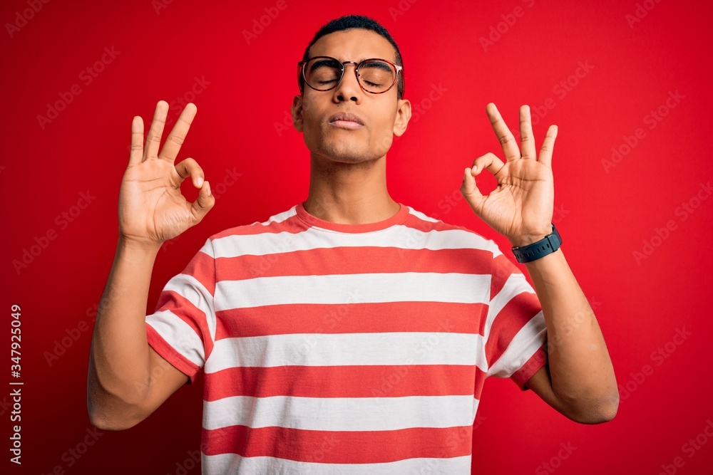 Young handsome african american man wearing casual striped t-shirt and glasses relaxed and smiling with eyes closed doing meditation gesture with fingers. Yoga concept. Young handsome african american man wearing casual striped t-shirt and glasses relaxed and smiling with eyes closed doing meditation gesture with fingers. Yoga concept.