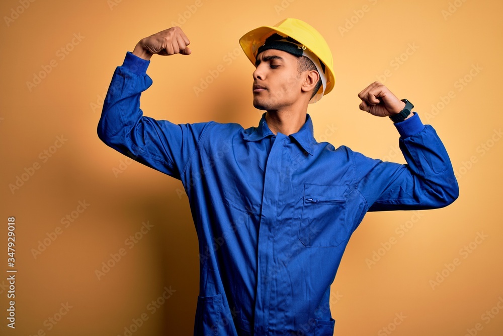 Young handsome african american worker man wearing blue uniform and security helmet showing arms muscles smiling proud. Fitness concept.