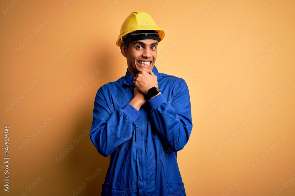 Young handsome african american worker man wearing blue uniform and security helmet laughing nervous and excited with hands on chin looking to the side