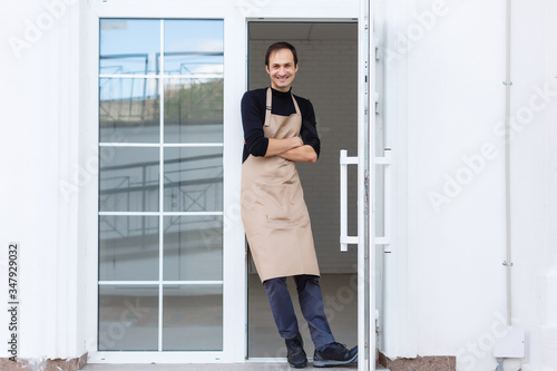 Portrait of smiling young man with facial hair wearing apron and leaning against bar counter in cafe. Blank slate board copy space for text or image.Cozy coffee shop with bartender owner staff service