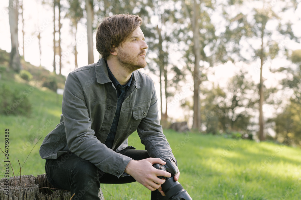 Portrait of young man sitting on tree stump outside holding camera Portrait of young man sitting on tree stump outside holding camera