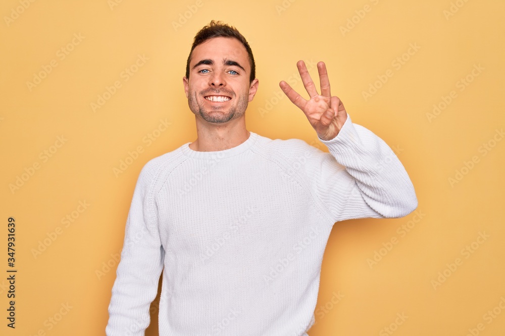 Young handsome man with blue eyes wearing casual sweater standing over yellow background showing and pointing up with fingers number three while smiling confident and happy. Young handsome man with blue eyes wearing casual sweater standing over yellow background showing and pointing up with fingers number three while smiling confident and happy.