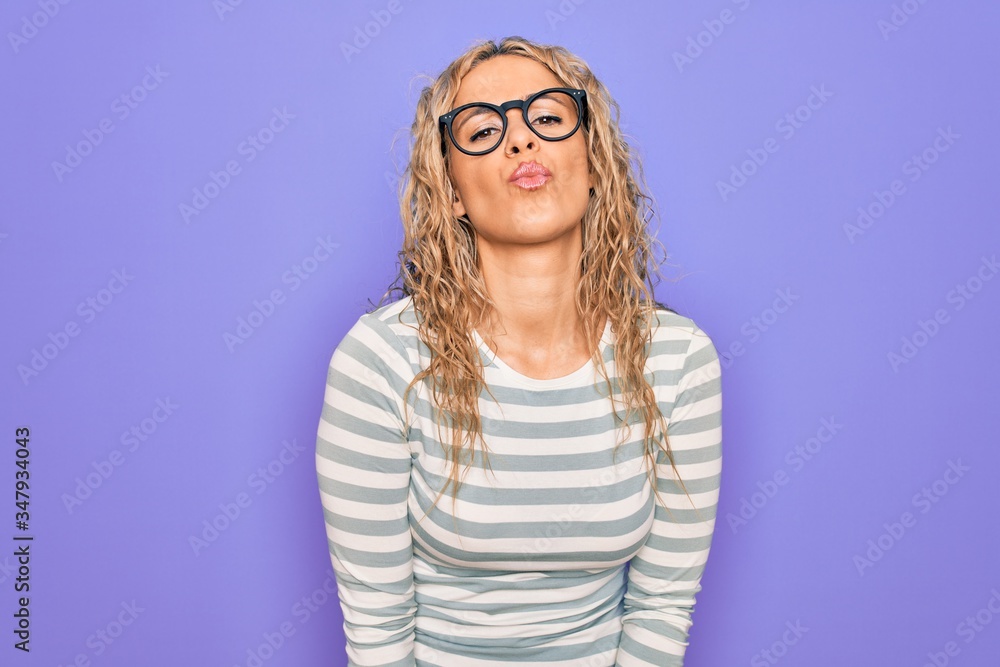 Naklejka premium Beautiful blonde woman wearing casual striped t-shirt and glasses over purple background looking at the camera blowing a kiss on air being lovely and sexy. Love expression.