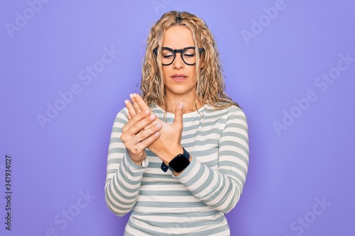 Beautiful blonde woman wearing casual striped t-shirt and glasses over purple background Suffering pain on hands and fingers, arthritis inflammation
