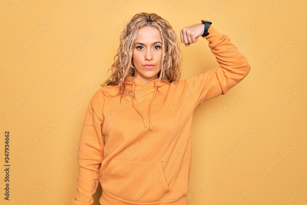Young beautiful blonde sporty woman wearing casual sweatshirt over yellow background Strong person showing arm muscle, confident and proud of power Young beautiful blonde sporty woman wearing casual sweatshirt over yellow background Strong person showing arm muscle, confident and proud of power