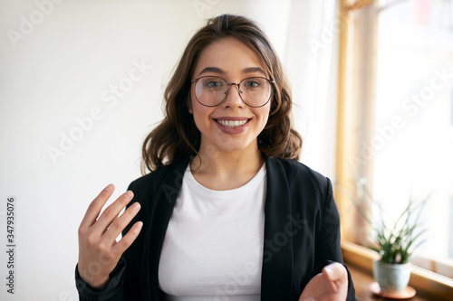 Beautiful positive young woman blogger recording course or workshop for her followers. Cute girl in eyewear chatting from home making video call using webcam and wirelesshigh speed internet connection photo
