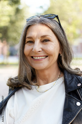 Fotografie Close up vertical shot of trendy looking middle aged sixty year old Caucasian female enjoying nice walk in park on sunny day, looking at camera with broad carefree happy smile