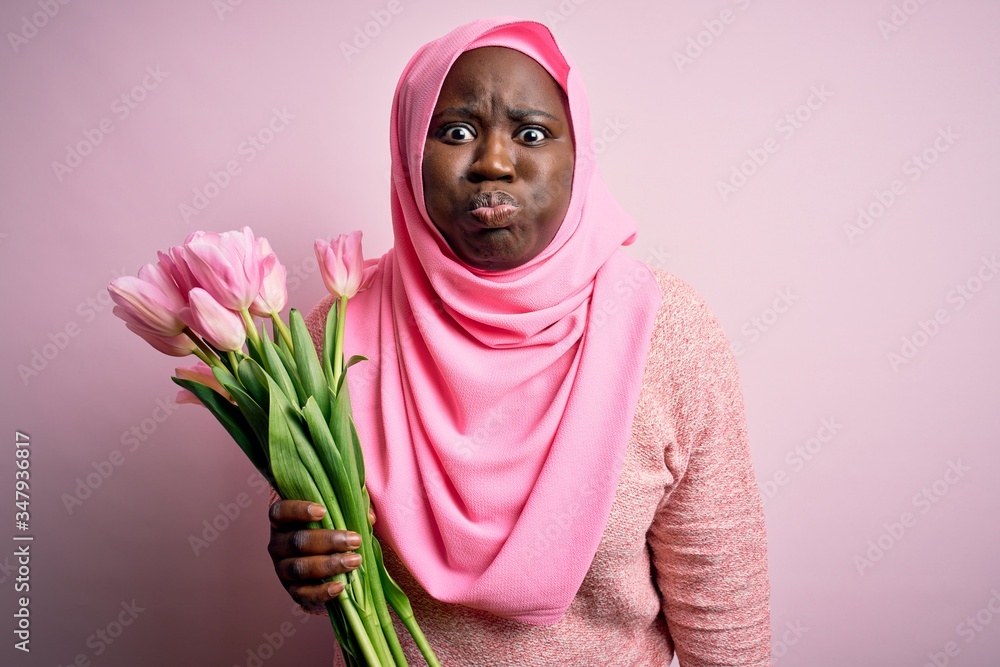 Young african american plus size woman wearing muslim hijab holding bouquet of pink tulips puffing cheeks with funny face. Mouth inflated with air, crazy expression. Young african american plus size woman wearing muslim hijab holding bouquet of pink tulips puffing cheeks with funny face. Mouth inflated with air, crazy expression.