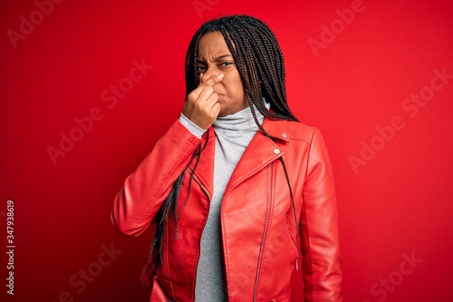 Young african american woman wearing cool fashion leather jacket over red isolated background smelling something stinky and disgusting, intolerable smell, holding breath with fingers on nose. Bad