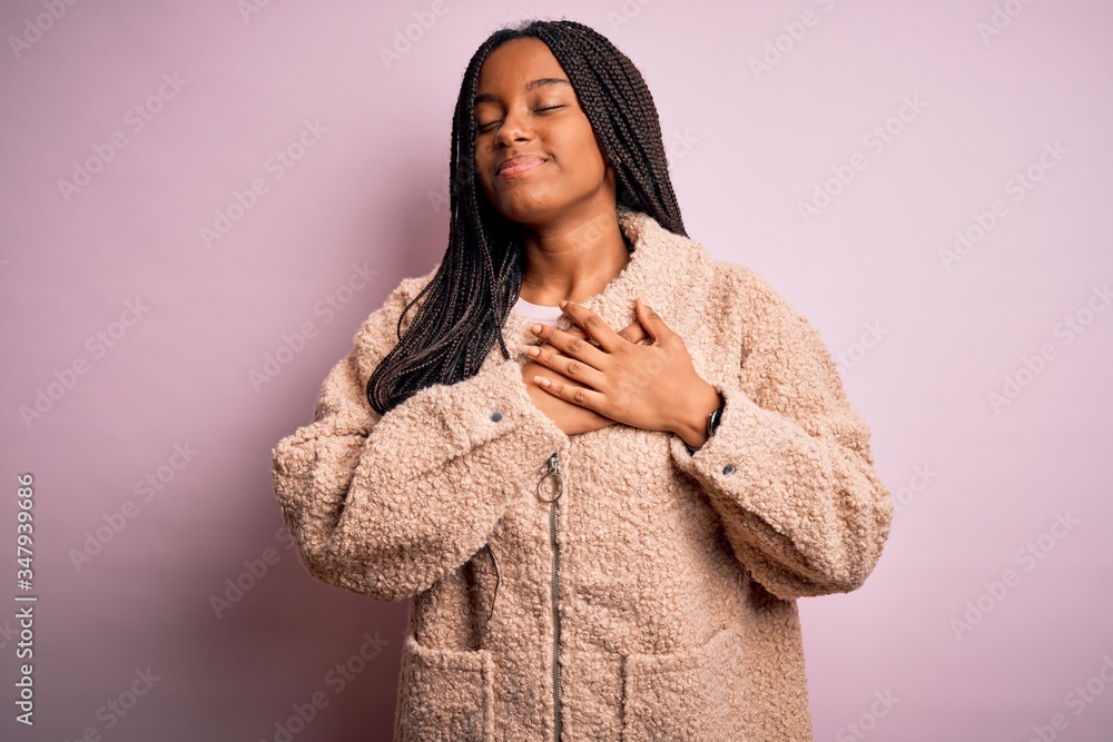 Young african american woman wearing fashion winter coat over pink isolated background smiling with hands on chest with closed eyes and grateful gesture on face. Health concept. Young african american woman wearing fashion winter coat over pink isolated background smiling with hands on chest with closed eyes and grateful gesture on face. Health concept.