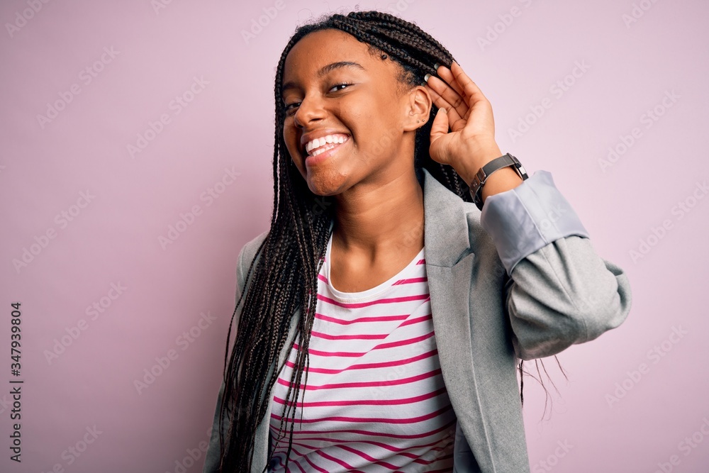 Young african american business woman standing over pink isolated background smiling with hand over ear listening an hearing to rumor or gossip. Deafness concept.