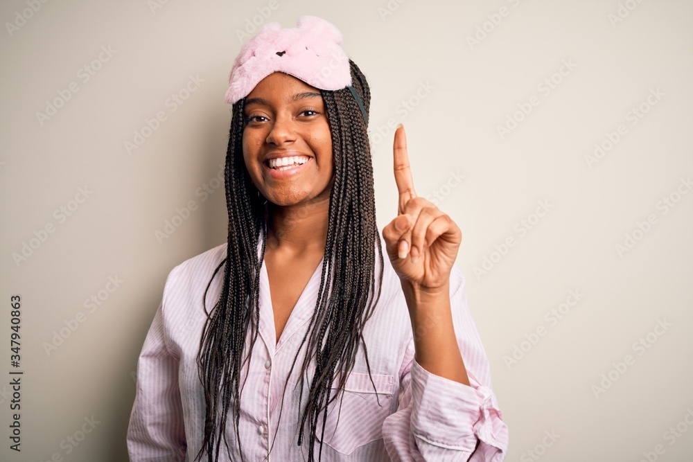 Young african american woman wearing pink pajama and sleep mask over isolated background showing and pointing up with finger number one while smiling confident and happy. Young african american woman wearing pink pajama and sleep mask over isolated background showing and pointing up with finger number one while smiling confident and happy.