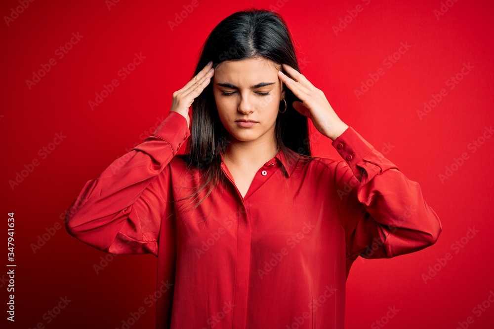 Young beautiful brunette woman wearing casual shirt standing over red background with hand on head for pain in head because stress. Suffering migraine. Young beautiful brunette woman wearing casual shirt standing over red background with hand on head for pain in head because stress. Suffering migraine.