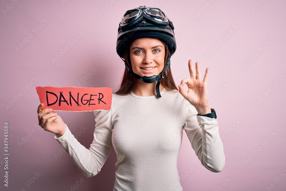 Beautiful motorcyclist woman with blue eyes wearing moto helmet holding danger message doing ok sign with fingers, excellent symbol Beautiful motorcyclist woman with blue eyes wearing moto helmet holding danger message doing ok sign with fingers, excellent symbol