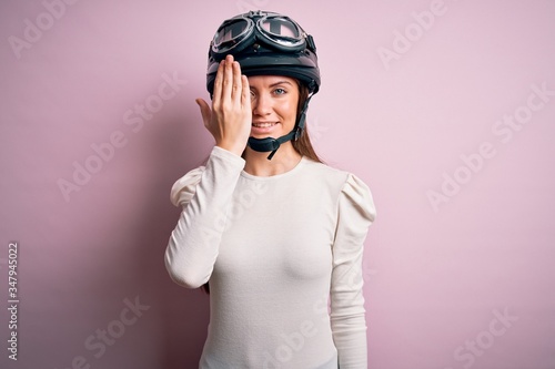 Young beautiful motorcyclist woman with blue eyes wearing moto helmet over pink background covering one eye with hand, confident smile on face and surprise emotion.
