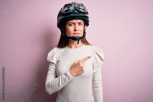 Young beautiful motorcyclist woman with blue eyes wearing moto helmet over pink background Pointing aside worried and nervous with forefinger, concerned and surprised expression