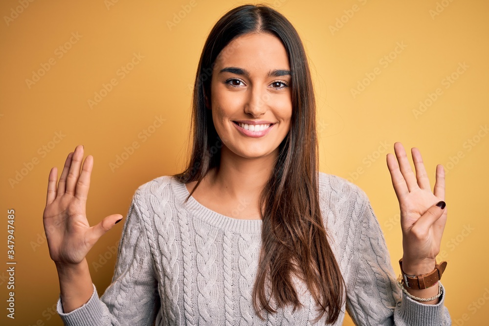 Young beautiful brunette woman wearing white casual sweater over yellow background showing and pointing up with fingers number nine while smiling confident and happy. Young beautiful brunette woman wearing white casual sweater over yellow background showing and pointing up with fingers number nine while smiling confident and happy.