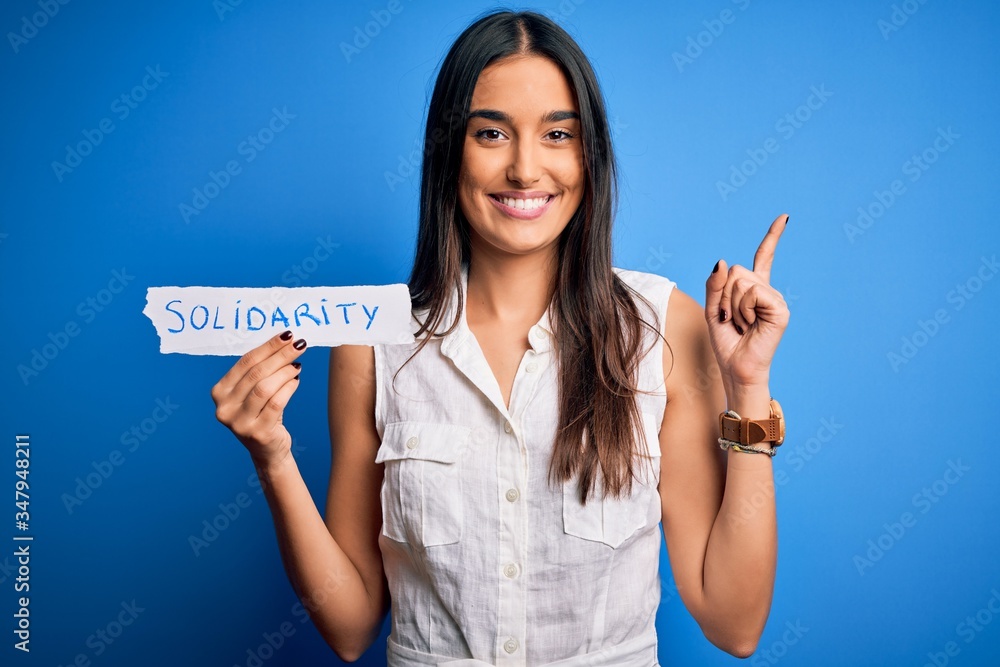 Young beautiful brunette woman holding paper with solidarity message over blue background surprised with an idea or question pointing finger with happy face, number one Young beautiful brunette woman holding paper with solidarity message over blue background surprised with an idea or question pointing finger with happy face, number one