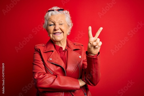 Obraz na plátně Senior beautiful grey-haired woman wearing casual red jacket and sunglasses smiling with happy face winking at the camera doing victory sign