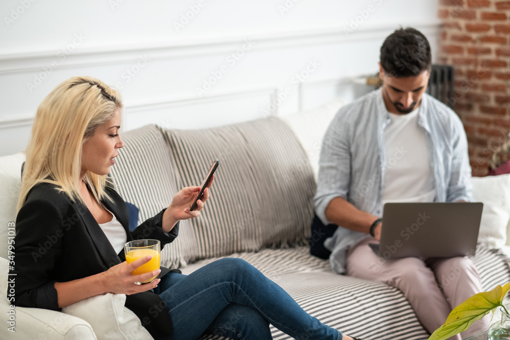 Positive blonde woman with cup of juice browsing smartphone and sitting on couch near ethnic boyfriend typing on laptop keyboard in living room of modern apartment Positive blonde woman with cup of juice browsing smartphone and sitting on couch near ethnic boyfriend typing on laptop keyboard in living room of modern apartment