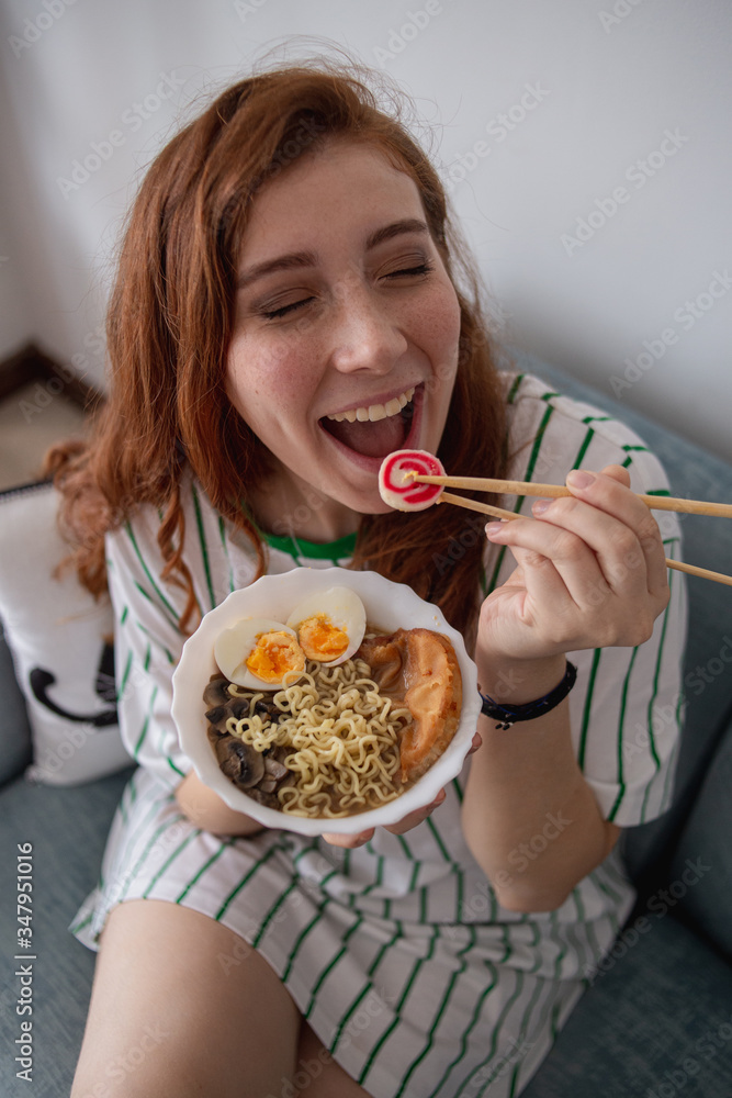 From above cheerful redhead female laughing and picking noodles from bowl of tasty ramen while sitting on couch at home From above cheerful redhead female laughing and picking noodles from bowl of tasty ramen while sitting on couch at home