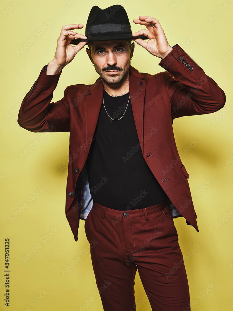 Handsome bearded well dressed male in trendy vinous suit and hat looking at camera against yellow background Handsome bearded well dressed male in trendy vinous suit and hat looking at camera against yellow background