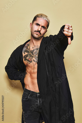 Young provocative male in black coat over naked tattooed torso showing thumbs down gesture in disapproval while standing against beige background photo