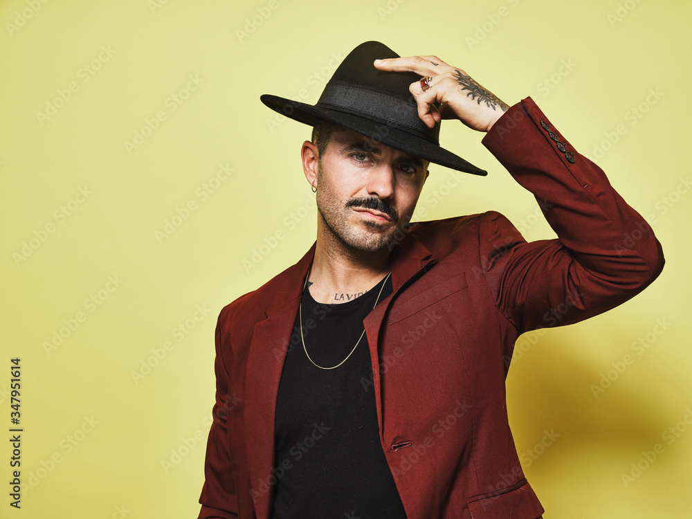 Handsome bearded well dressed male in trendy vinous suit and hat looking at camera against yellow background Handsome bearded well dressed male in trendy vinous suit and hat looking at camera against yellow background