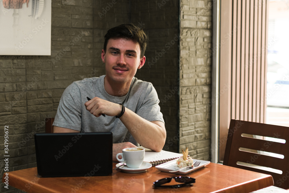 Young man with laptop and notebook smiling and looking at camera while sitting at table with coffee and salad in cozy restaurant Young man with laptop and notebook smiling and looking at camera while sitting at table with coffee and salad in cozy restaurant