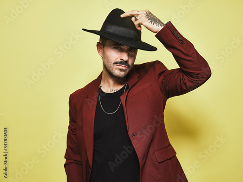 Handsome bearded well dressed male in trendy vinous suit and hat looking at camera against yellow background