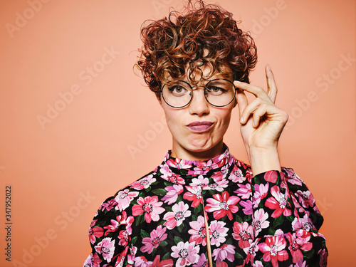 Thoughtful doubtful beautiful curly haired female in trendy eyeglasses and stylish colorful blouse with floral ornament looking at camera against pink background