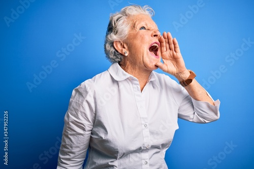Senior beautiful woman wearing elegant shirt standing over isolated blue background shouting and screaming loud to side with hand on mouth. Communication concept.
