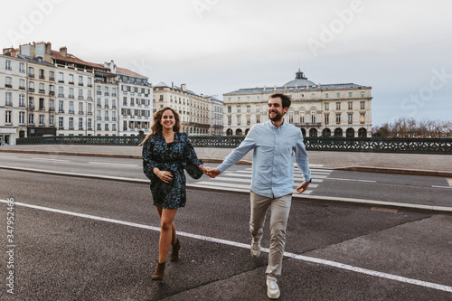 Happy young romantic couple in stylish clothes laughing and holding hands while crossing bridge with historic buildings in background during city tour in Bayonne in France
