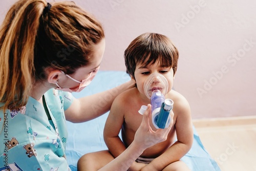 Side view of young female pediatric doctor in medical mask doing inhalation treatment with nebulizer for little boy having respiratory problem in hospital photo