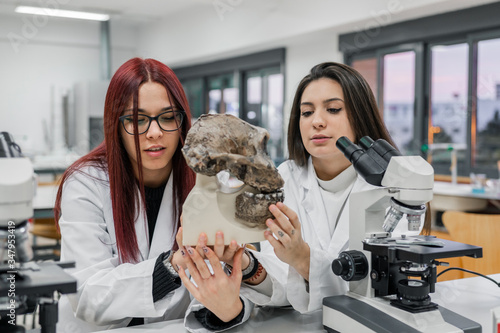 Young women in white robes examining old skull of primate near microscope during anthropology lesson in contemporary laboratory photo