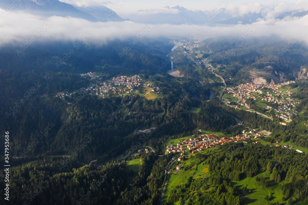 Italian Dolomites mountain valley with town in the country. Aerial beautiful shot above the clouds. Green hills with buildings. Tiled roofs on the houses. Fields with forests.