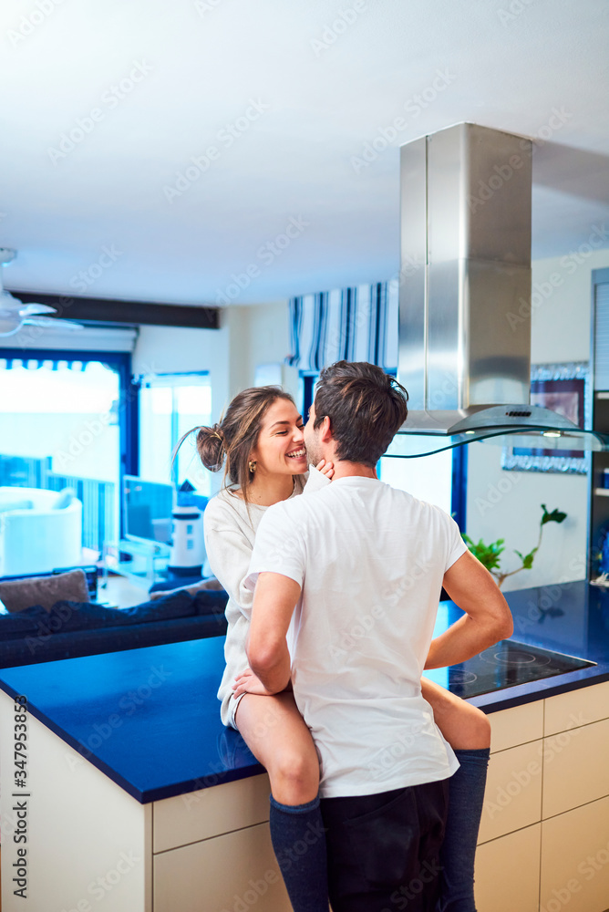 Side view of happy young woman sitting on kitchen counter and embracing loving husband while spending day together in modern apartment Side view of happy young woman sitting on kitchen counter and embracing loving husband while spending day together in modern apartment