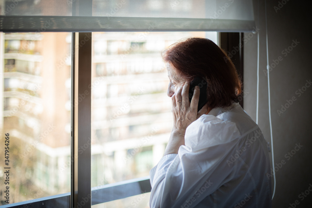 Side view of senior female in white blouse having phone call while standing near window in hospital ward visiting relative Side view of senior female in white blouse having phone call while standing near window in hospital ward visiting relative
