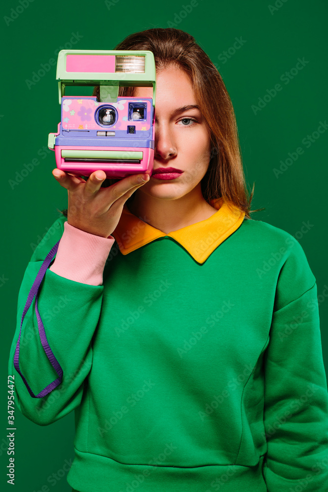 Young unemotional female in green pullover taking picture with retro instant camera while standing against green background