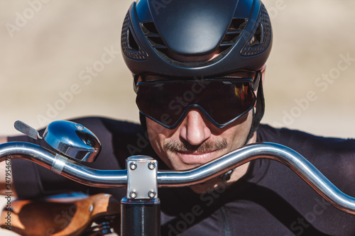 Portrait of happy man in black activewear and helmet looking at camera while standing with retro penny farthing bicycle on racetrack at sports ground