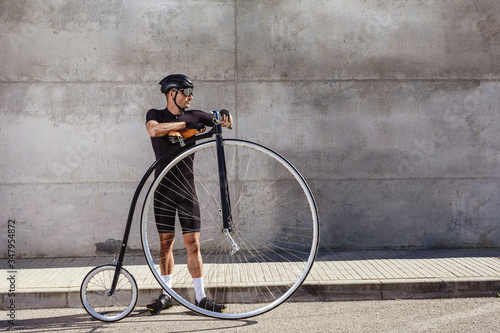 Calm determined man in black activewear and helmet looking away while standing on asphalt road and leaning on high wheel bicycle against concrete wall in sunny day photo