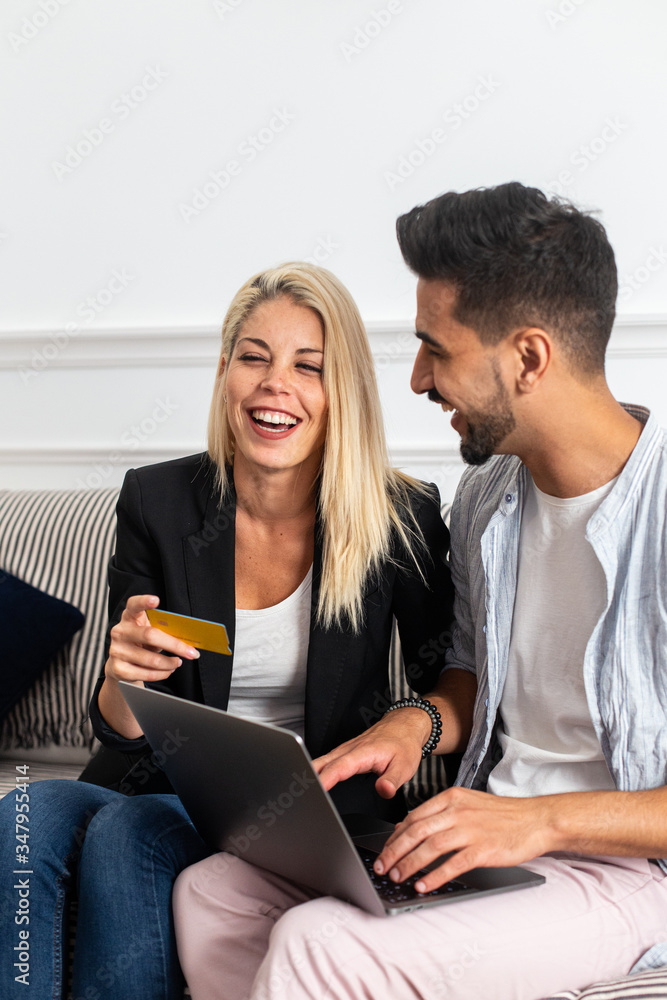 Delighted blond female smiling and reading credit card credentials to cheerful ethnic boyfriend with laptop while sitting on sofa and making online purchases together Delighted blond female smiling and reading credit card credentials to cheerful ethnic boyfriend with laptop while sitting on sofa and making online purchases together