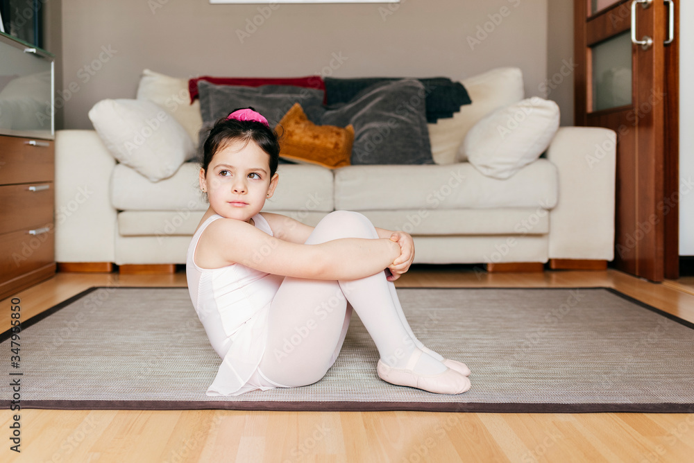 Bored thoughtful little girl in leotard and tights sitting on floor looking  away while resting during ballet rehearsal at home Stock Photo