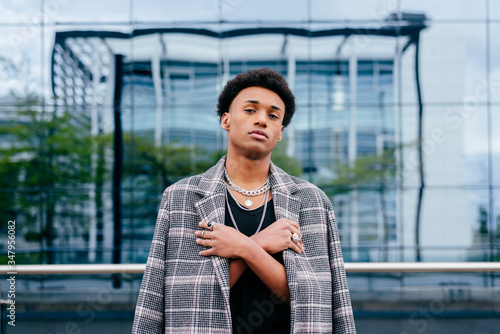 Confident young African American male teenager model in trendy tartan coat and stylish accessories looking at camera while standing against contemporary building with glass wall photo