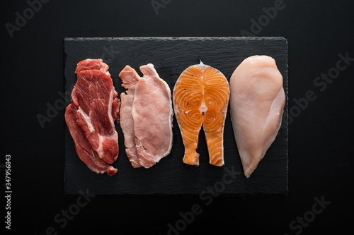 From above fresh raw fillet of beef and pork with chicken and fish pieces on wooden cutting board as composition of protein rich food against black background photo