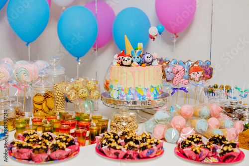 delicious and beautiful cake on a birthday table with many different sweets, gingerbread cookies, marshmallows and colored balloons