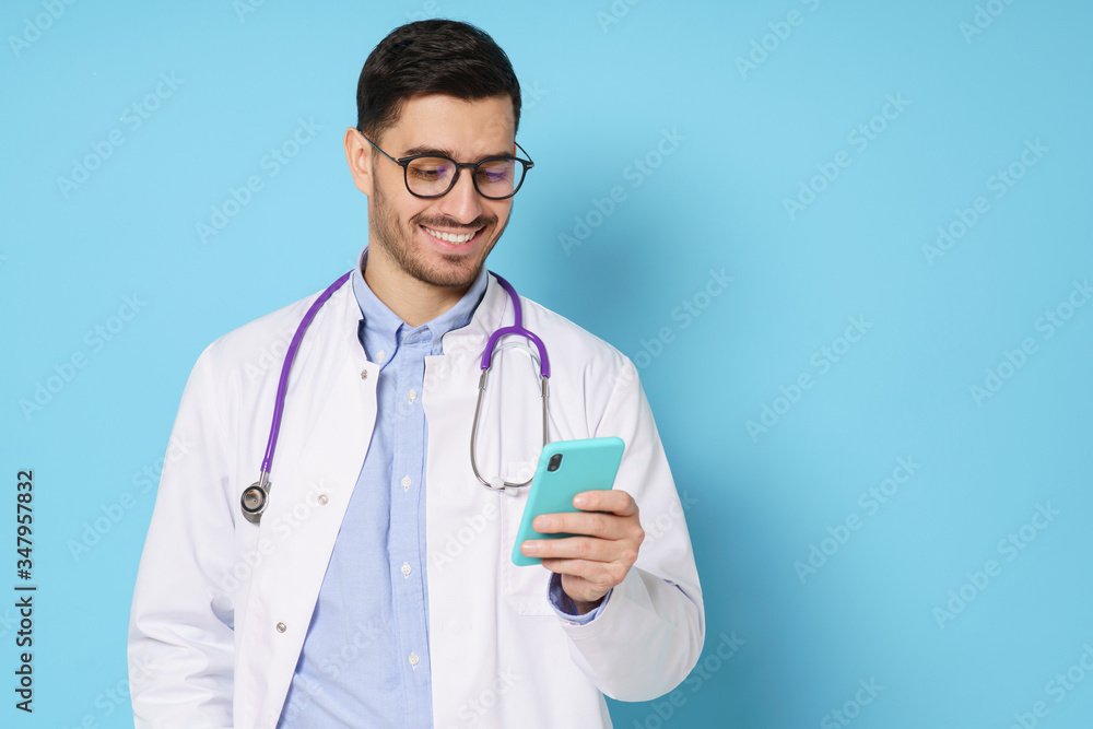Young male doctor looking at smart phone screen with smile, exchanging messages with colleagues, isolated on blue background, copy space on right Young male doctor looking at smart phone screen with smile, exchanging messages with colleagues, isolated on blue background, copy space on right