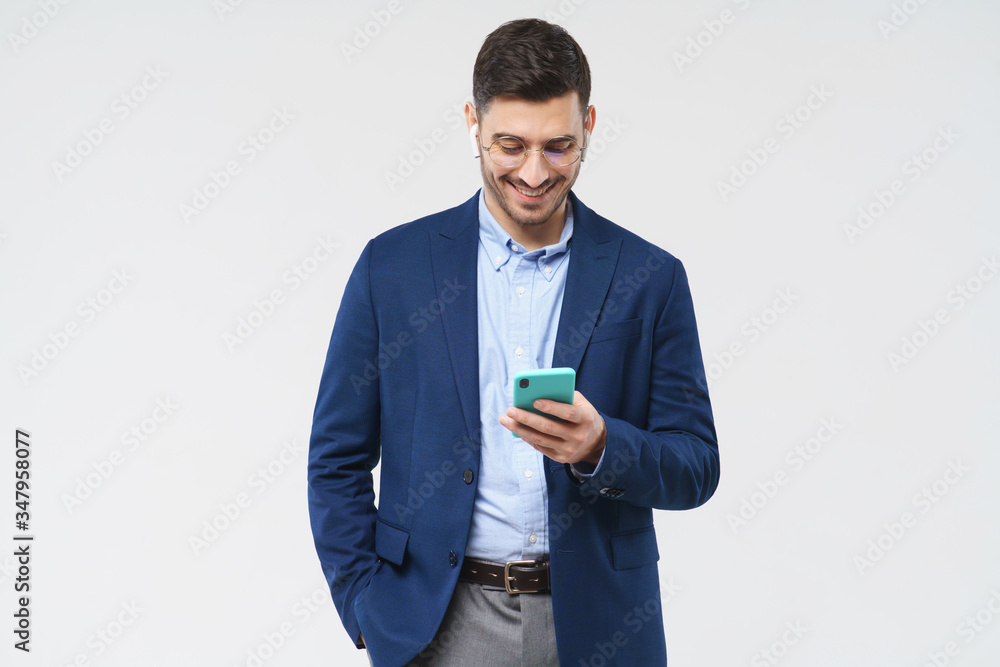 Portrait of young male dressed in formal clothes, wearing glasses, holding phone in one hand, browsing newsfeed with smile, isolated on gray background Portrait of young male dressed in formal clothes, wearing glasses, holding phone in one hand, browsing newsfeed with smile, isolated on gray background