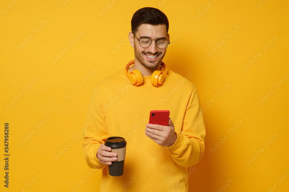 Millennial man in glasses, enjoying messages from friends via phone screen, smiling happily, drinking coffee, headphones round neck, isolated on yellow background Millennial man in glasses, enjoying messages from friends via phone screen, smiling happily, drinking coffee, headphones round neck, isolated on yellow background